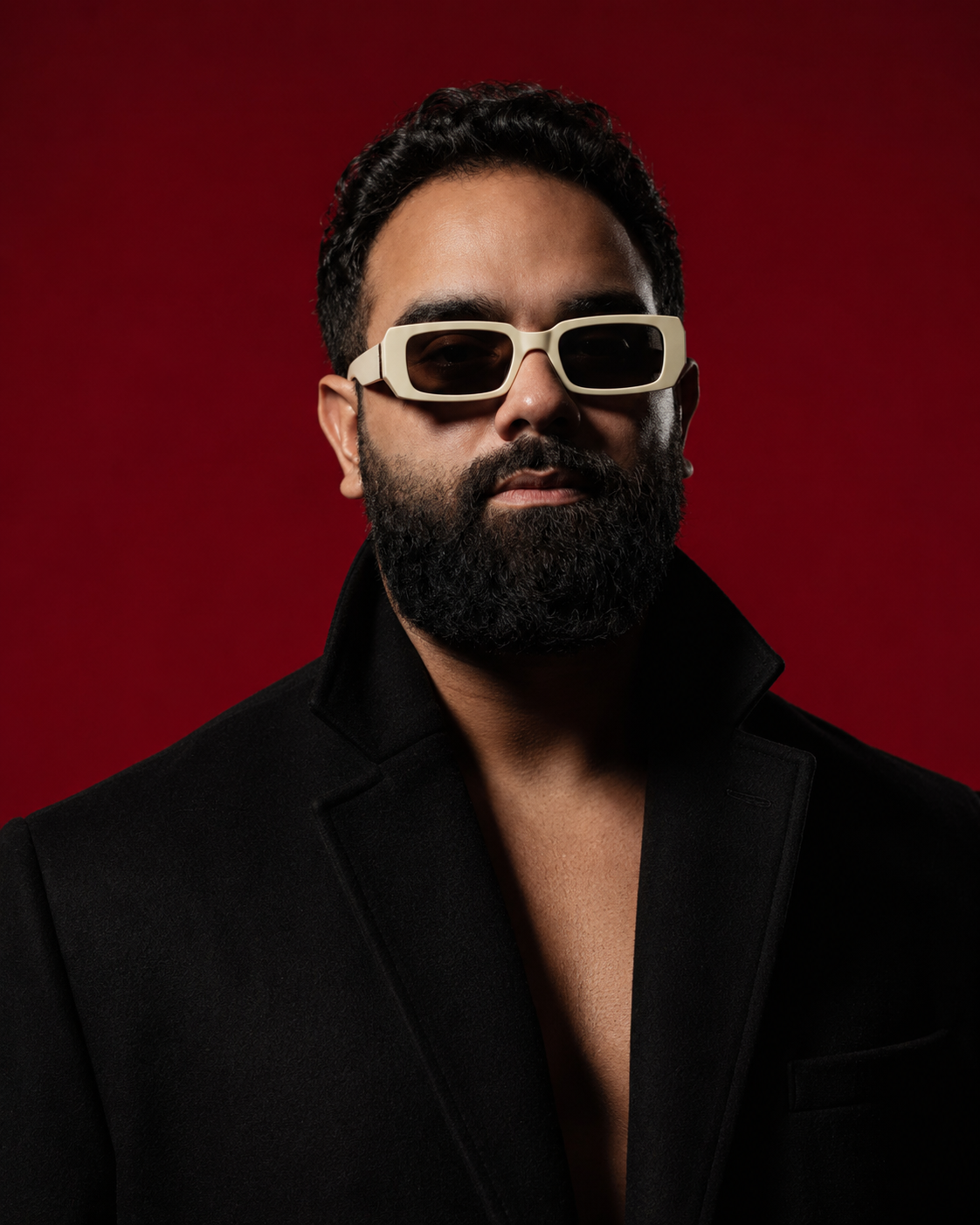 Editorial portrait of a bearded man wearing bold cream rectangular sunglasses and a black tailored coat with an open neckline, posed against a deep red studio background. Dramatic directional lighting creates strong shadows and a sleek, high-fashion atmos