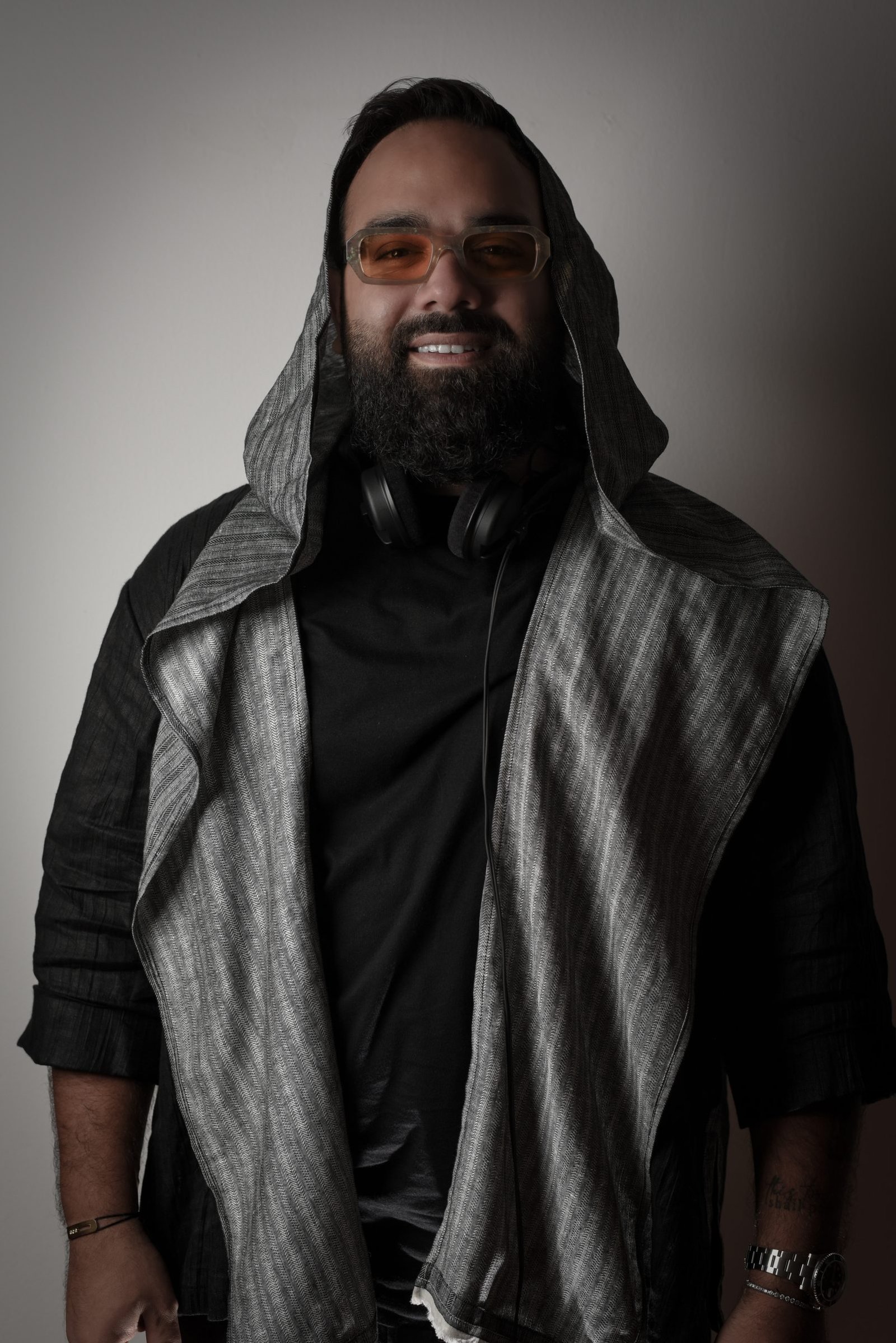 DJ LEMIR smiling with the hood of a striped overshirt up, wearing amber-tinted glasses, headphones around the neck.